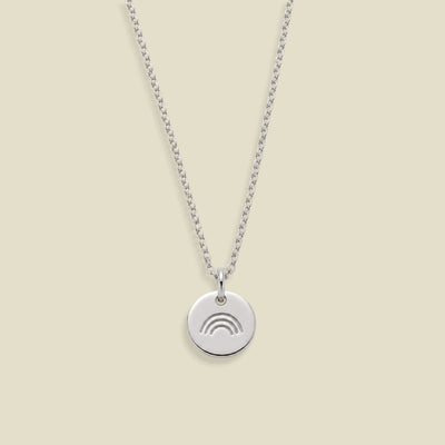 A silver chain necklace with a small round disc pendant featuring a minimalist rainbow engraving, displayed against a solid light beige background.