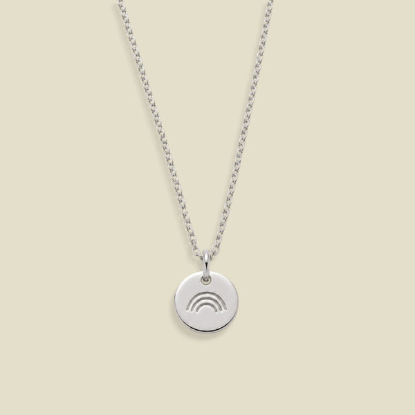 A silver chain necklace with a small round disc pendant featuring a minimalist rainbow engraving, displayed against a solid light beige background.