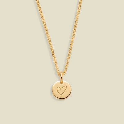 A gold chain necklace with a small round disc pendant engraved with a heart, displayed against a solid light beige background.