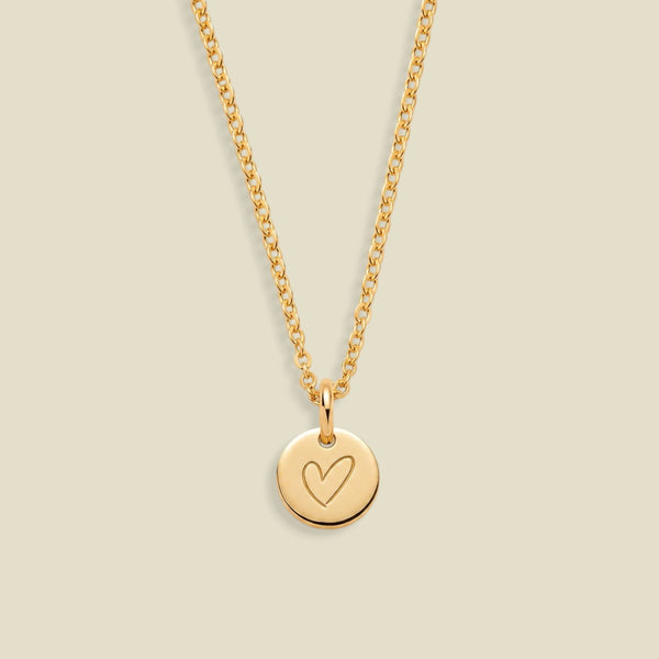 A gold chain necklace with a small round disc pendant engraved with a heart, displayed against a solid light beige background.