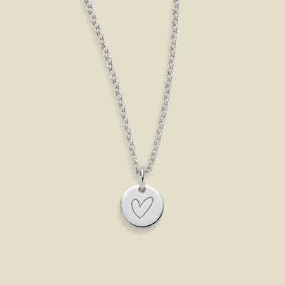 A silver chain necklace with a round silver disc pendant engraved with a heart, displayed against a solid light beige background.