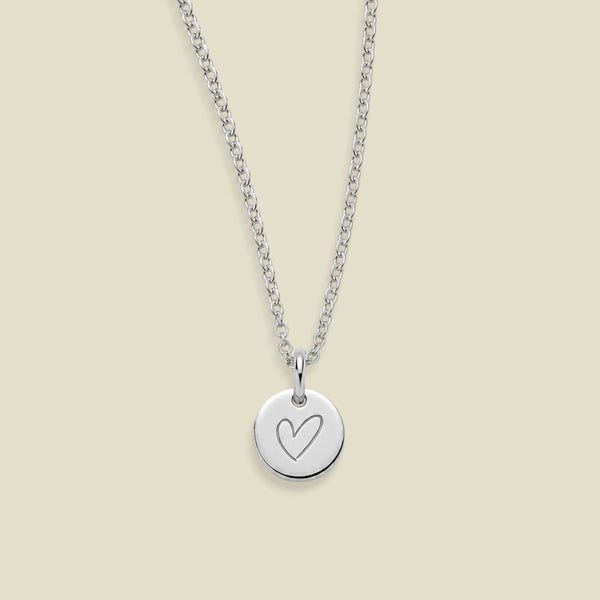 A silver chain necklace with a round silver disc pendant engraved with a heart, displayed against a solid light beige background.