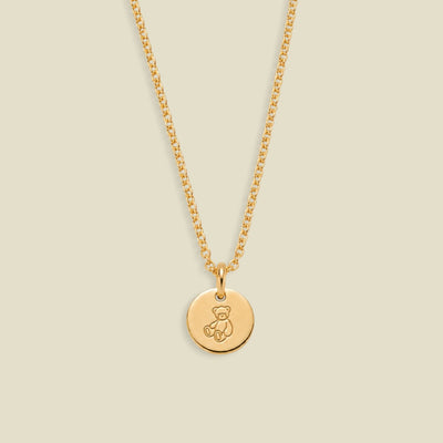 A gold chain necklace with a round disc pendant engraved with a teddy bear, shown against a solid light beige background.