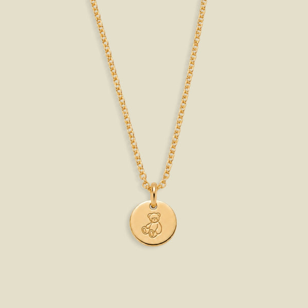 A gold chain necklace with a round disc pendant engraved with a teddy bear, shown against a solid light beige background.