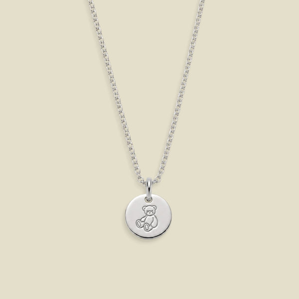 A silver round disc pendant necklace featuring an engraved teddy bear design, suspended from a delicate silver chain against a solid light beige background.