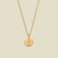 A delicate gold chain necklace with a small round gold disc pendant featuring an engraved cactus symbol, displayed against a solid light beige background.