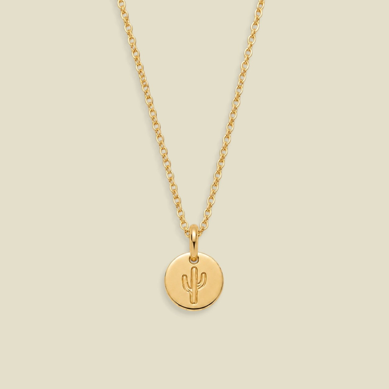 A delicate gold chain necklace with a small round gold disc pendant featuring an engraved cactus symbol, displayed against a solid light beige background.