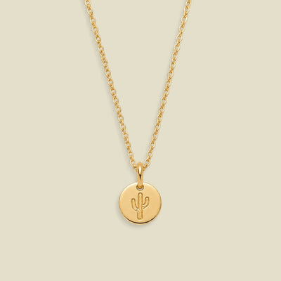 A delicate gold chain necklace with a small round gold disc pendant featuring an engraved cactus symbol, displayed against a solid light beige background.
