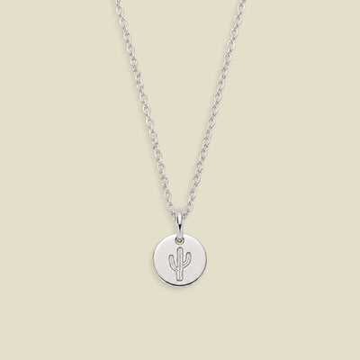 A silver chain necklace with a round silver disc pendant engraved with a cactus, displayed against a solid light beige background.