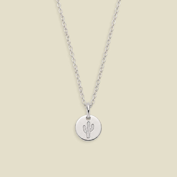 A silver chain necklace with a round silver disc pendant engraved with a cactus, displayed against a solid light beige background.
