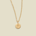 A gold chain necklace with a small round disc pendant engraved with a palm tree, displayed against a solid light beige background.