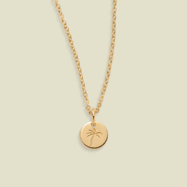 A gold chain necklace with a small round disc pendant engraved with a palm tree, displayed against a solid light beige background.