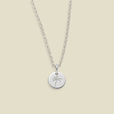 A silver chain necklace with a round disc pendant engraved with a palm tree, displayed against a solid beige background.