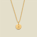A gold chain necklace with a round gold disc pendant engraved with a small pine tree, displayed against a solid light beige background.