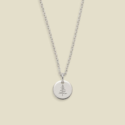 A silver chain necklace with a round silver disc pendant engraved with a small pine tree, displayed against a solid light beige background.
