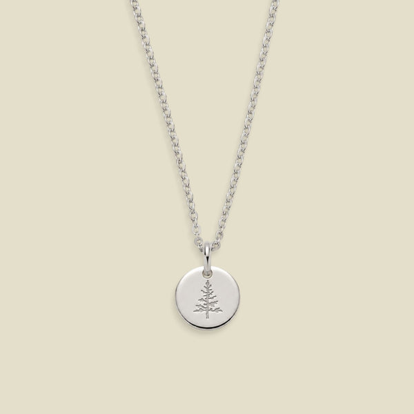 A silver chain necklace with a round silver disc pendant engraved with a small pine tree, displayed against a solid light beige background.