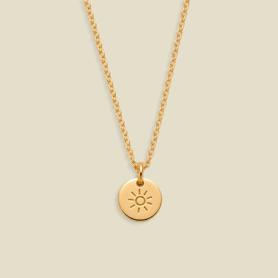 A gold chain necklace with a small round disc pendant featuring an engraved sun design, displayed against a solid light beige background.