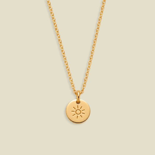 A gold chain necklace with a small round disc pendant featuring an engraved sun design, displayed against a solid light beige background.