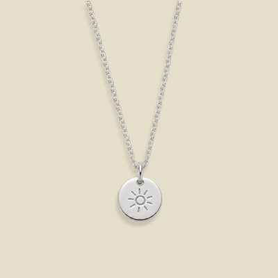 A silver chain necklace with a small round disc pendant featuring an engraved sun design, displayed against a solid light beige background.