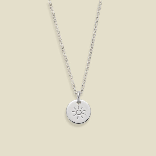 A silver chain necklace with a small round disc pendant featuring an engraved sun design, displayed against a solid light beige background.