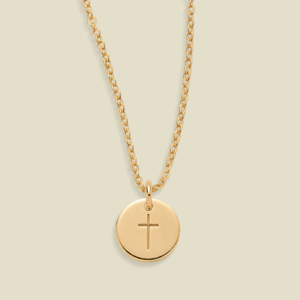 A gold chain necklace with a round gold disc pendant engraved with a cross, displayed against a solid light beige background.