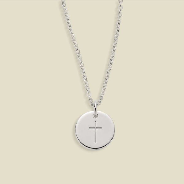A silver chain necklace with a round silver disc pendant engraved with a cross, displayed against a solid light beige background.