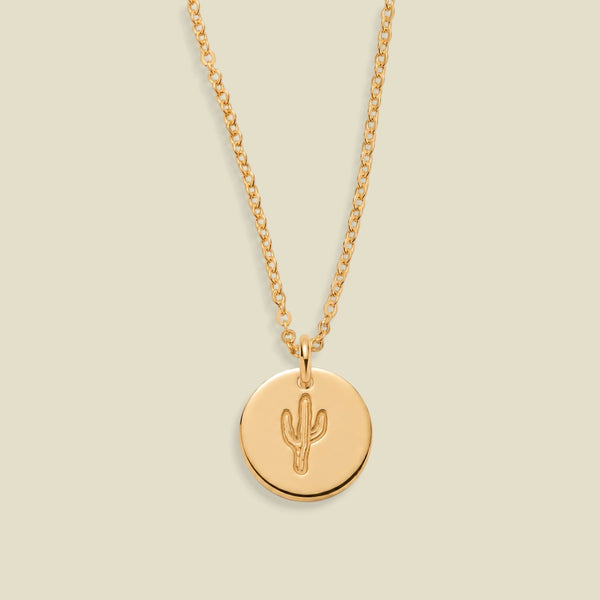 A gold chain necklace with a round gold pendant featuring an engraved cactus design, displayed against a solid light beige background.