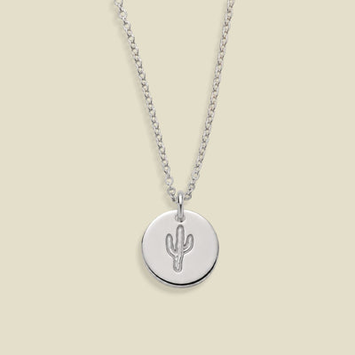 A silver chain necklace with a round silver disc pendant featuring a simple cactus design, displayed against a solid light beige background.