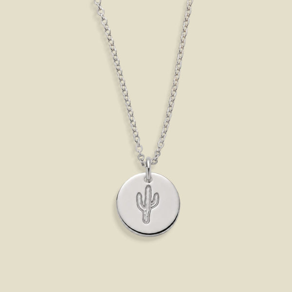 A silver chain necklace with a round silver disc pendant featuring a simple cactus design, displayed against a solid light beige background.