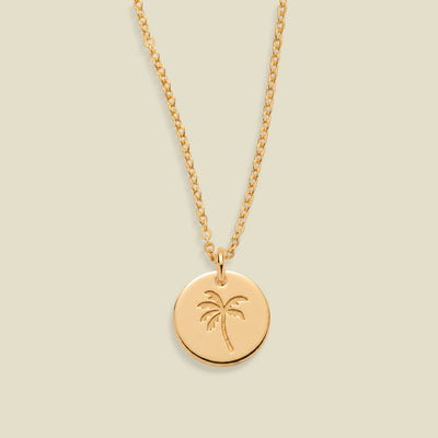 A gold chain necklace with a round gold disc pendant engraved with a palm tree, shown against a solid light beige background.