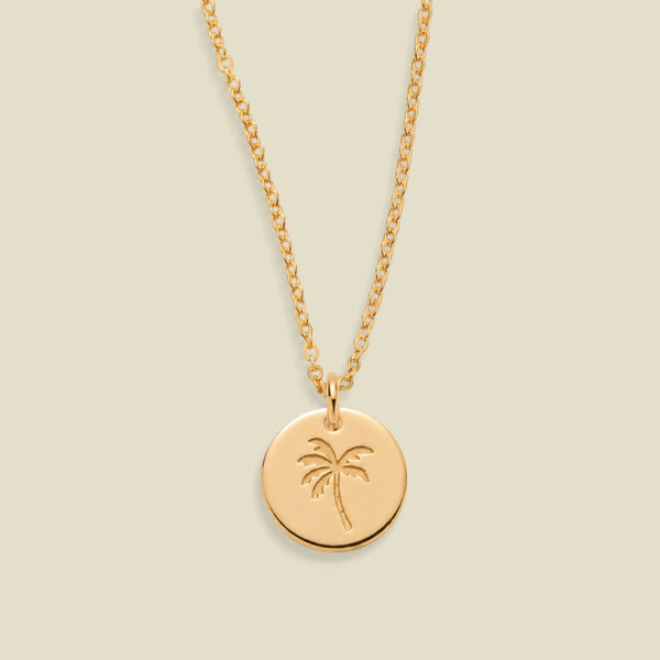 A gold chain necklace with a round gold disc pendant engraved with a palm tree, shown against a solid light beige background.