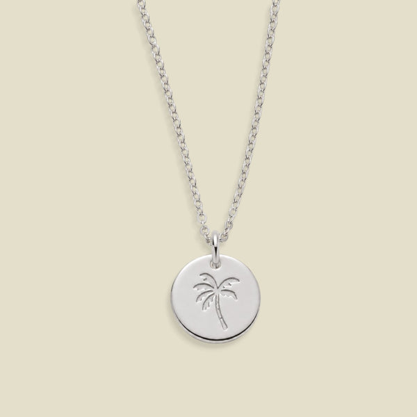 A silver round disc pendant necklace featuring an engraved palm tree design, suspended from a silver chain against a solid light beige background.