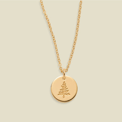A gold chain necklace with a round gold disc pendant engraved with a small pine tree, displayed against a solid light beige background.