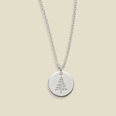 A silver round disc pendant necklace featuring an engraved pine tree design, suspended from a delicate silver chain against a solid light beige background.