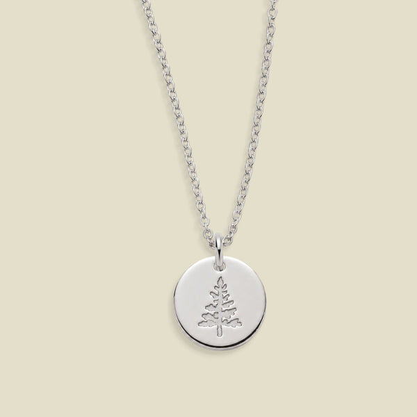 A silver round disc pendant necklace featuring an engraved pine tree design, suspended from a delicate silver chain against a solid light beige background.