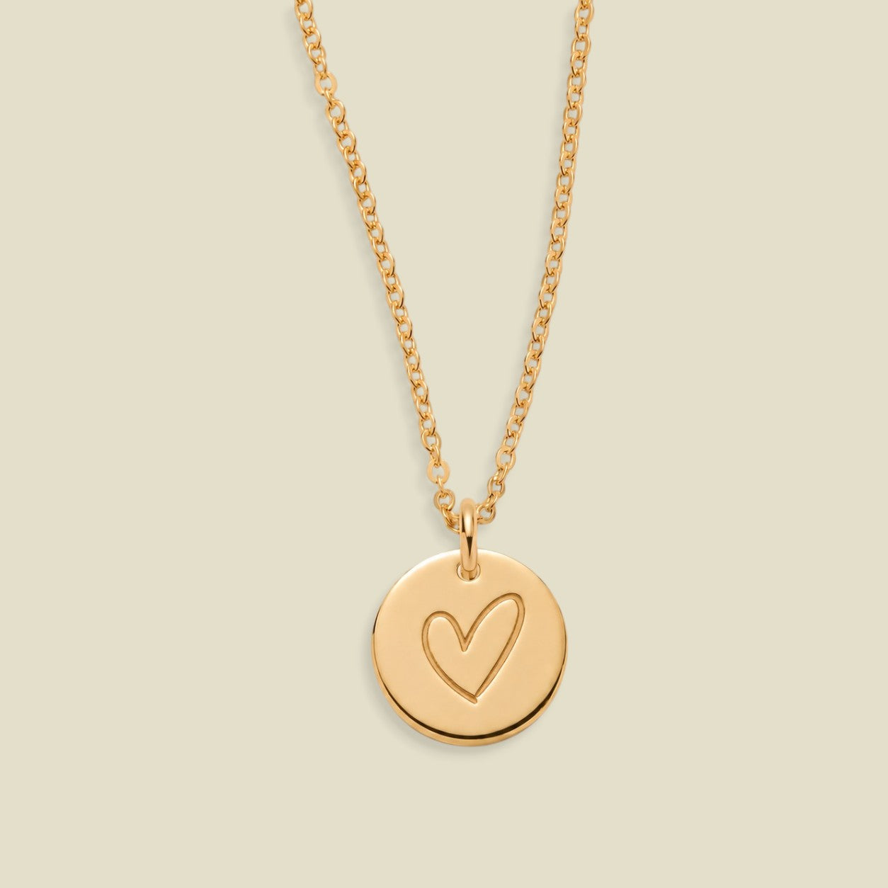 A gold chain necklace with a round gold disc pendant engraved with a heart, displayed against a solid light beige background.