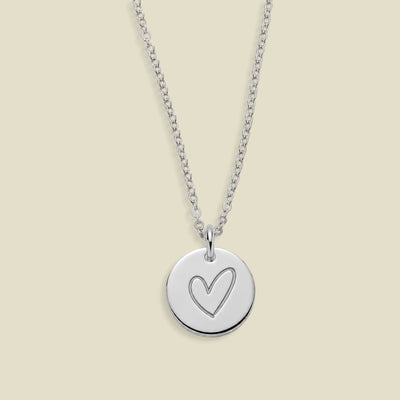 A silver round disc pendant necklace with a simple heart outline engraving, suspended from a delicate silver cable chain against a solid light beige background.