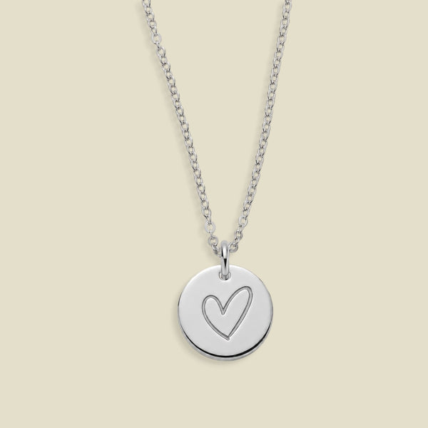 A silver round disc pendant necklace with a simple heart outline engraving, suspended from a delicate silver cable chain against a solid light beige background.
