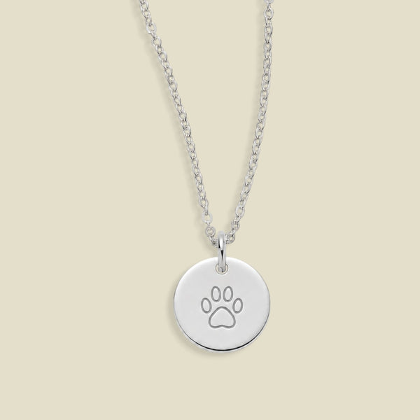 A silver round disc pendant necklace engraved with a paw print, hanging from a delicate silver chain against a solid light beige background.