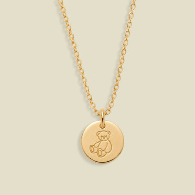 A gold chain necklace with a round disc pendant engraved with a teddy bear, shown against a solid light beige background.