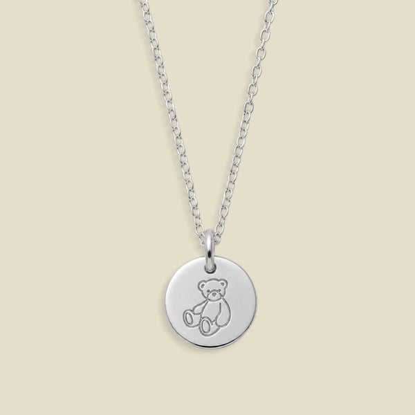 A silver round disc pendant necklace featuring an engraved teddy bear design, suspended from a delicate silver chain against a solid light beige background.