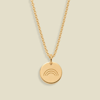A gold chain necklace with a round gold disc pendant featuring an engraved rainbow design, displayed against a solid light beige background.