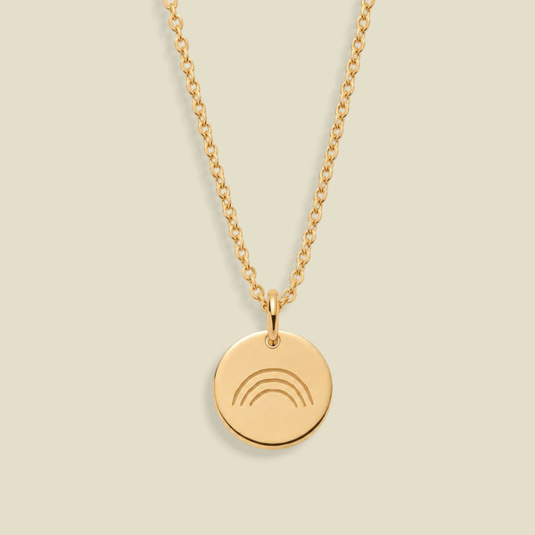A gold chain necklace with a round gold disc pendant featuring an engraved rainbow design, displayed against a solid light beige background.