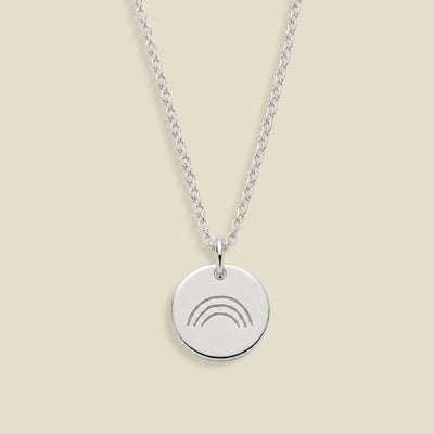 A silver chain necklace with a round silver pendant engraved with a simple rainbow design, displayed against a solid light beige background.
