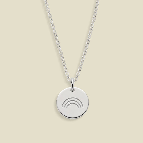 A silver chain necklace with a round silver pendant engraved with a simple rainbow design, displayed against a solid light beige background.