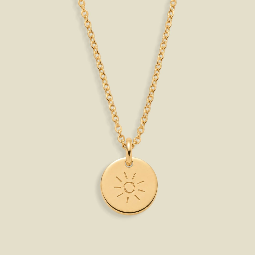 A gold chain necklace with a round gold pendant engraved with a sun face, displayed against a solid light beige background.