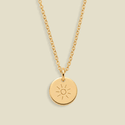 A gold chain necklace with a round gold pendant engraved with a sun face, displayed against a solid light beige background.