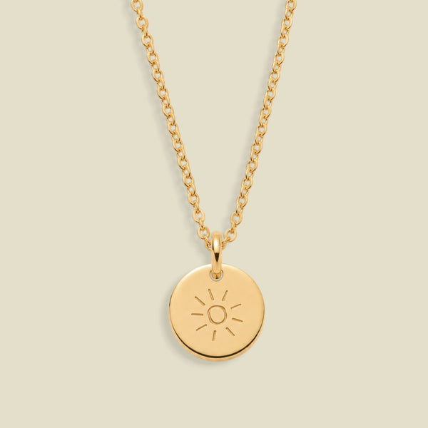 A gold chain necklace with a round gold pendant engraved with a sun face, displayed against a solid light beige background.