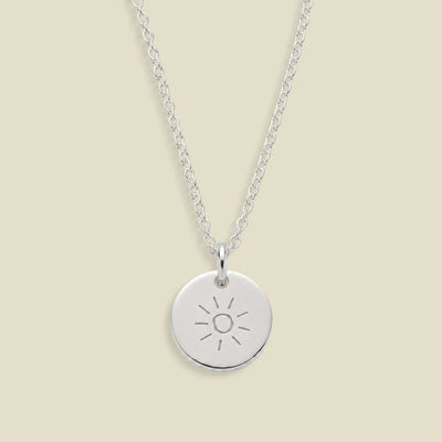 A silver round disc pendant necklace engraved with a sun design, hanging from a delicate silver chain against a solid light beige background.