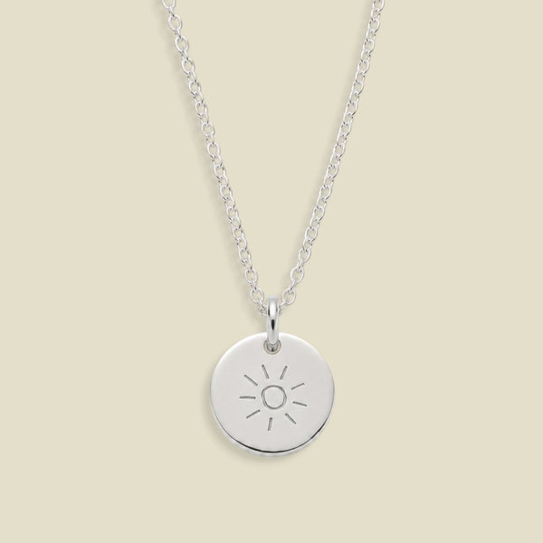 A silver round disc pendant necklace engraved with a sun design, hanging from a delicate silver chain against a solid light beige background.